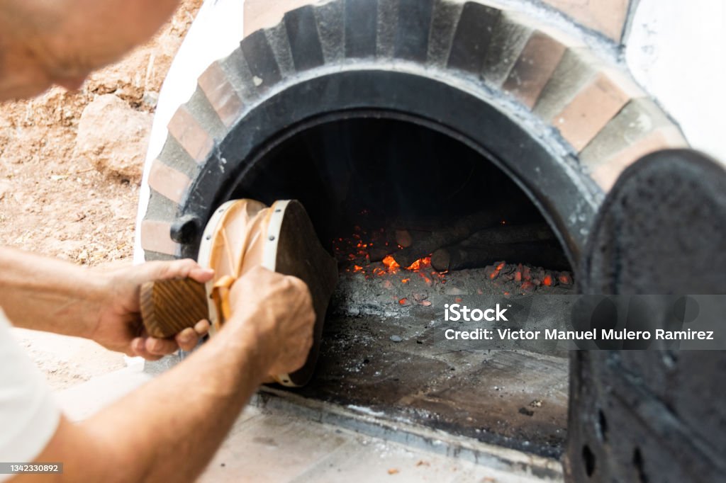 Artisan wood-fired oven and man's hands blowing the embers with a bellows White painted artisan wood-fired oven built on the outside with the door open and man's hands blowing the embers with a bellows Bellows Stock Photo