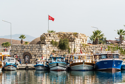 Sigacik, Izmir, Turkey - October 5, 2020. Crumbling 16th-century Genoese castle in Sigacik neighborhood of Seferihisar district of Izmir Province in Turkey. View from across the water, with fishing boats and people.