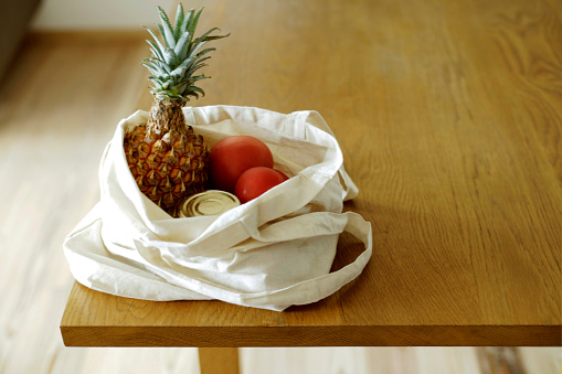 Off-white cloth bag with tomatoes, a pineapple and a can in it sitting on wooden dining table. Sunlit room. Space for copy.