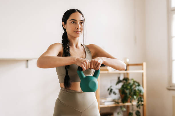 Gorgeous woman working-out with a kettlebell in her living room A sporty woman is exercising with a green kettlebell at home. Her eyes are focused. She is dressed in matching fashionable sports leggings and top. Furniture with a plant is visible behind her. Horizontal daylight photo. kettlebell stock pictures, royalty-free photos & images