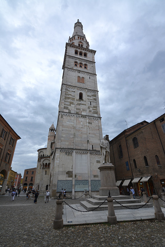 People walking in Piazza Grande by the Torre della Ghirlandina or Ghirlandina, the bell tower of the Cathedral of Modena, in Emilia-Romagna, Italy.\nStanding at 86.12 metres, the tower is the traditional symbol of Modena, being visible from all directions outside the city, Emilia Romagna.