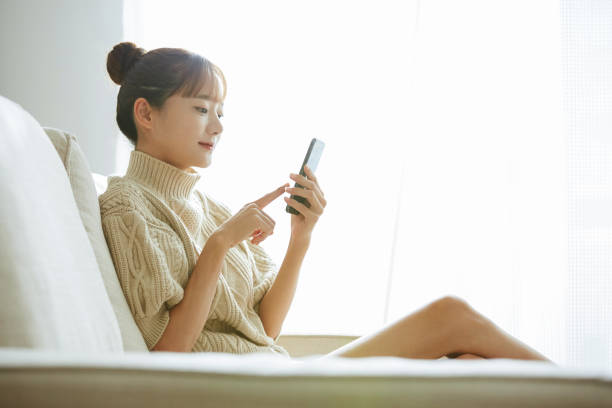自宅で電話を操作している若いアジアの女性 - スマホ 日本人 ストックフォトと画像