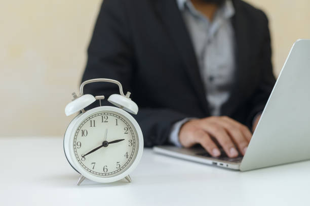 istock Analog white retro alarm clock on office table with businessman using laptop. working time concept. 1342391454