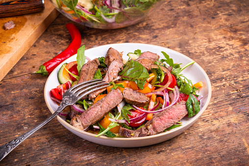Salad of vegetables and lettuce leaves with slices of medium-well steak on a plate with a fork - rustic style