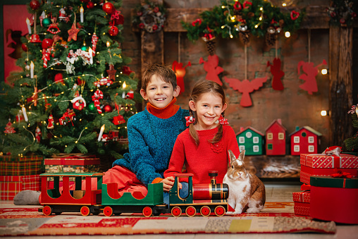 Cheerful brother and sister with a cute rabbit in Christmas play with a children's wooden train. Christmas mood.
