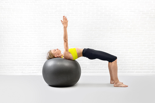 Slim athletic caucasian woman practice shoulder bridge drill using big exercise ball in fitness studio indoor, arms up. Glutes and hips workout, pilates, balance, wellness, gymnastics concept.