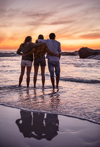 Rearview of a group of young friends standing arm in arm together in the ocean surf on a beach at sunset