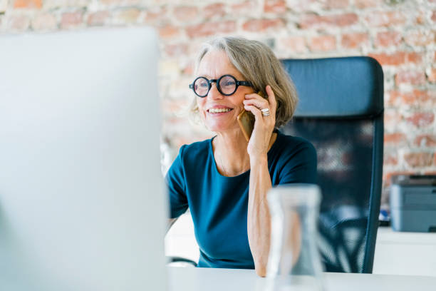 starsza bizneswoman przy biurku za pomocą telefonu komórkowego - computer computer monitor women business person zdjęcia i obrazy z banku zdjęć