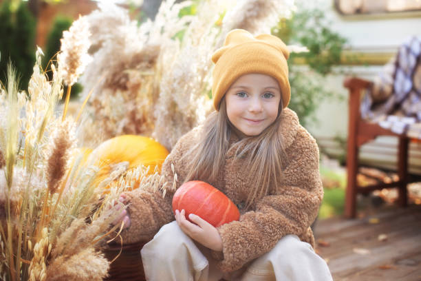 ハロウィーンで屋外カボチャと幸せな子供。笑顔の女の子は、彼女の手にカボチャを持つポーチに座っています。トリック・オア・トリート。秋の庭で家の近くで遊ぶ幸せな小さな女の子。� - single flower flower autumn pumpkin ストックフォトと画像
