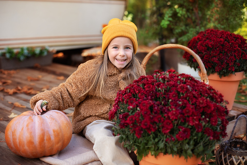 Happy little girl sitting on porch of house with chrysanthemum potted and pumpkins. home fall decoration for Halloween or Thanksgiving. Smilling Child in autumn garden with yellow pumpkins and flowers