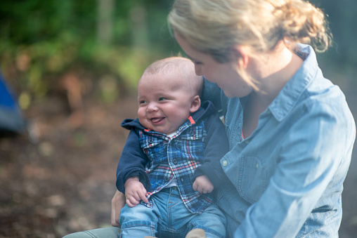 A mother sits with her infant son on her lap as they sit around a camp fire.  They are both dressed casually in layers as they watch the rest of their family roast marshmallows.  The mother is looking  down at the baby and smiling.