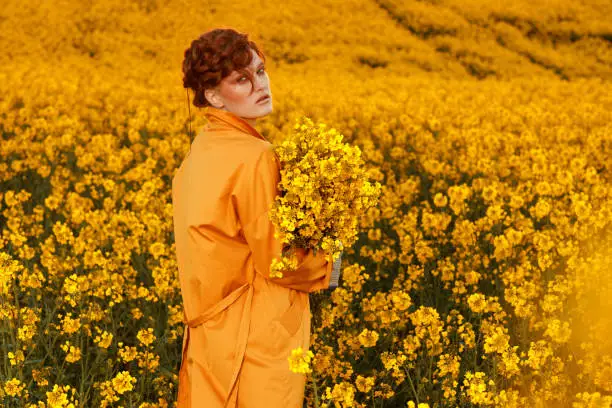 Photo of Portrait of red haired women dressed in trendy fall orange trench coat in colourful orange field. Fashion clothes autumn look