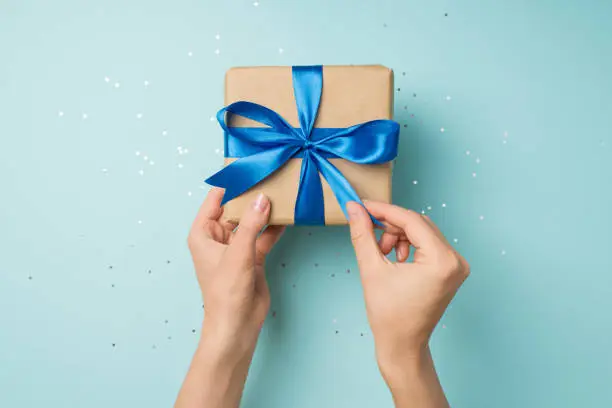 Photo of First person top view photo of hands unpacking craft paper giftbox with vivid blue satin ribbon bow over shiny sequins on isolated pastel blue background