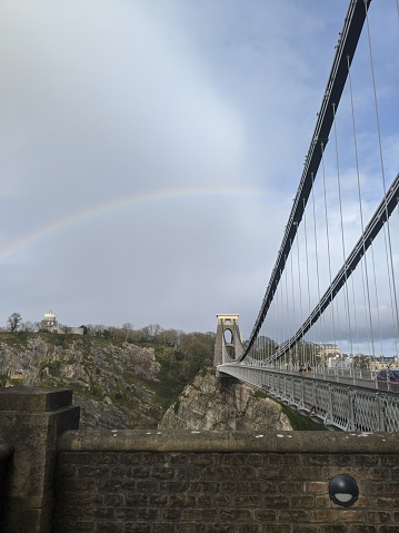An arched rainbow over the Clifton Suspension Bridge in Bristol. The sky is cloudy but the blue still shines through. .