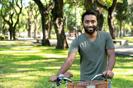 Cheerful Hindi young man taking a walk at the park next to his bicycle facing camera smiling - Lifestyles