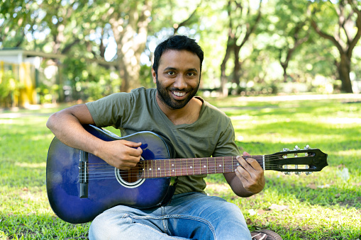 Young Hindi man enjoying a day at the park playing his guitar while smiling at camera very cheerfully - Lifestyles