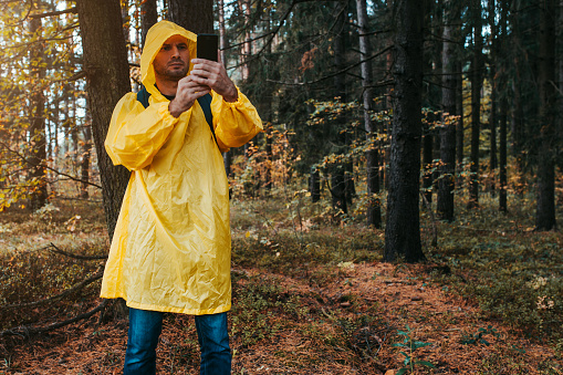 A man in a yellow raincoat takes pictures with a smartphone in a dark dense rainy forest