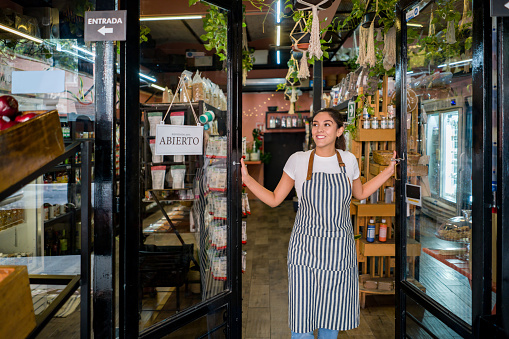 Happy Latin American woman opening the door at a supermarket - essential services concepts