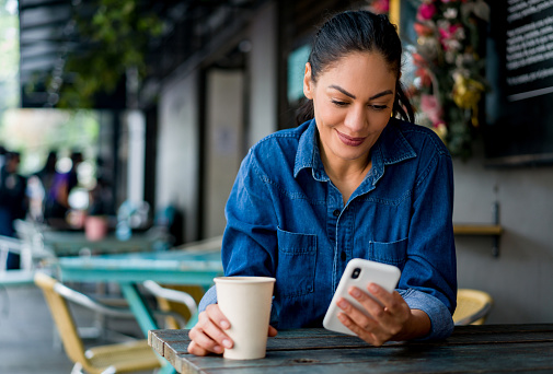 Portrait of a happy Latin American woman drinking a cup of coffee at a cafe while checking her cell phone and smiling - lifestyle concepts