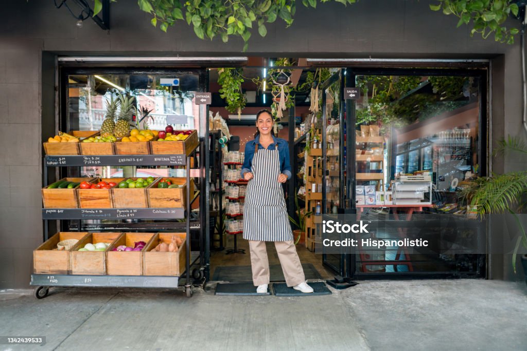Happy woman working at a local food market and standing at the door waiting for clients Happy Latin American woman working at a local food market and standing at the door waiting for clients - small business concepts Store Stock Photo