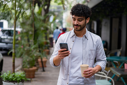 Portrait of a happy man on the go drinking a cup of coffee on the go while checking his cell phone on the street - lifestyle concepts