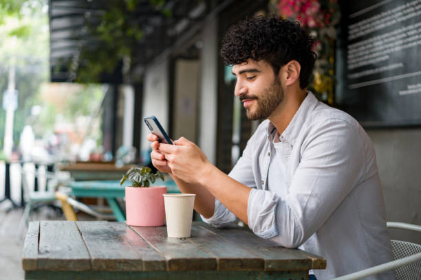 hombre feliz bebiendo revisando su teléfono celular en una cafeter�ía mientras bebe un capuchino - restaurant review fotografías e imágenes de stock