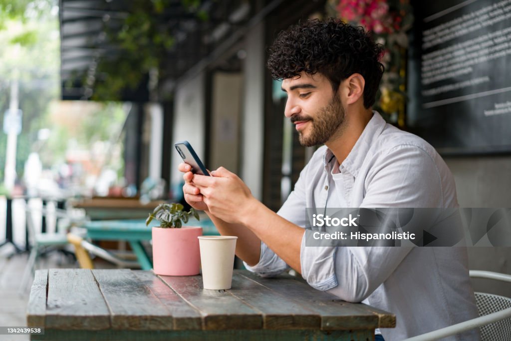 Happy man drinking checking his cell phone at a coffee shop while drinking a cappuccino Portrait of a happy Latin American man drinking checking his cell phone at a coffee shop while drinking a cappuccino - lifestyle concepts Men Stock Photo