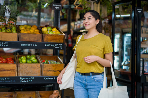 Happy Latin American woman shopping at a local food market using reusable bags and leaving the store smiling
