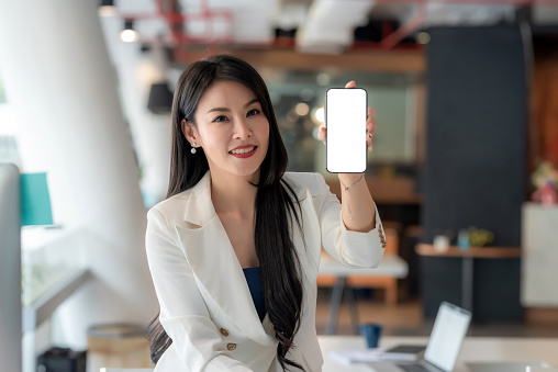 Beautiful Asian businesswoman looking at the camera holding a smartphone  blank white screen at the office. Mock up.
