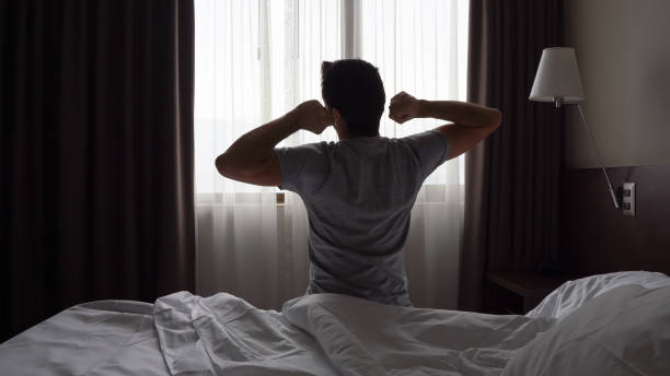 Silhouette of man sitting on bed outstretching by bedroom window Silhouette of man sitting on bed outstretching by bedroom window. Early morning wake up, lazy, tired, lack of energy concepts waking up stock pictures, royalty-free photos & images