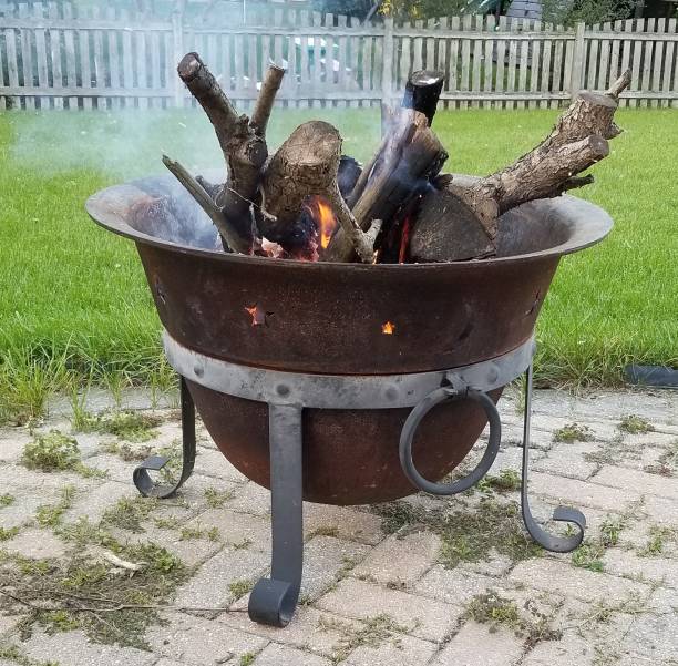 Side profile view of an old weathered and rusty large fire pit cauldron filled with firewood. Wood is burning and flames and smoke are visible. The iron kettle sits in a metal stand atop a weed covered brick patio pavement. Green grass and a wooden picket fence are in the background. Daytime on a crisp early Autumn afternoon.