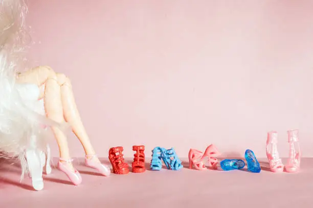 An articulated doll sitting in a chair four pairs of toy shoes in a row on a pink background