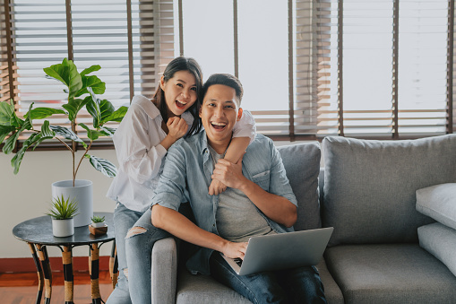 Happy Asian couple using laptop on sofa at home. Woman embracing man from behind