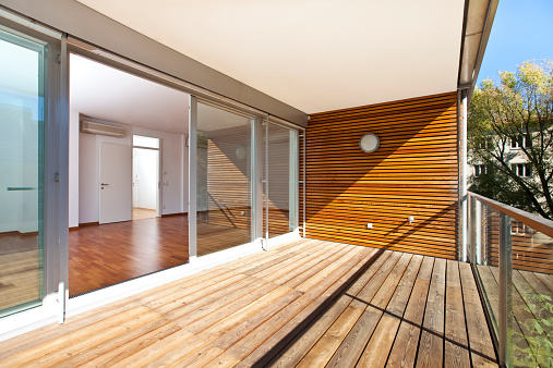sunlit balcony with wooden floor and wall of an architectural contemporarily apartment building in green area.