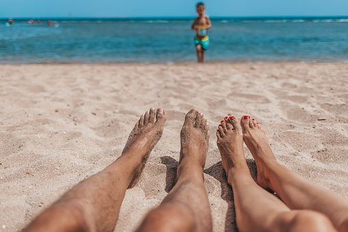 Sandy toes and feet of a couple enjoying a beach vacation while watching their kid playing in the sand.