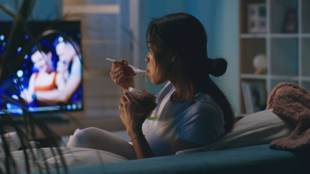 Female eating in front of TV Asian woman eating takeaway noodles and watching TV while sitting on couch and having dinner in evening at home watching tv stock pictures, royalty-free photos & images