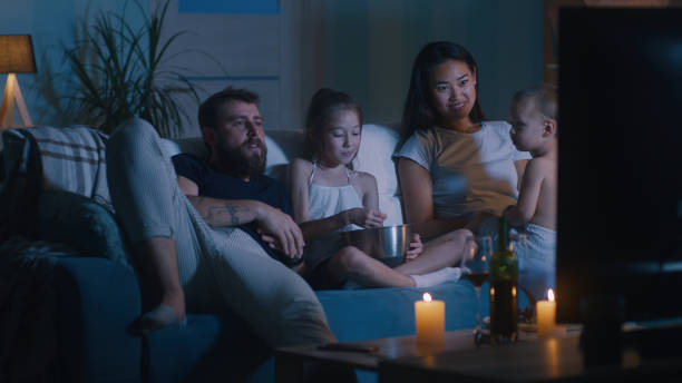 Diverse family watching movie in dark living room Bearded man throwing popcorn into mouth while chilling on sofa and watching movie with wife and kids in dark living room at night at home asian kids watching tv stock pictures, royalty-free photos & images