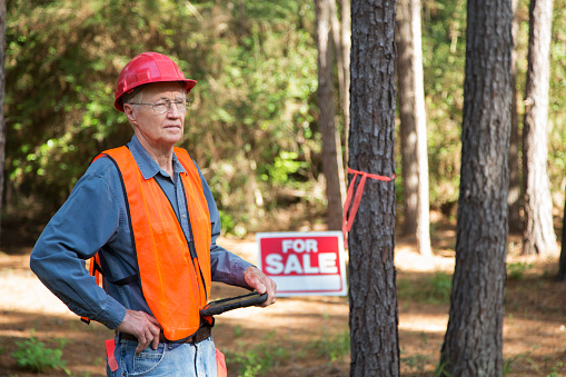 Construction manager marking trees for be cut.  He carries a digital tablet and wears a safety vest and hard hat.  For sale sign is on the premises.