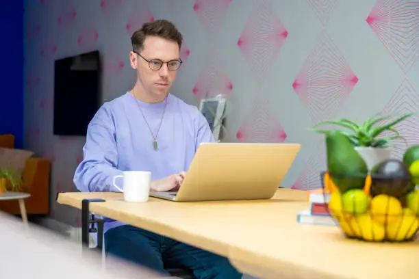 Casually dressed Caucasian man with short brown hair and eyeglasses sitting at dining table with mug of coffee and typing on laptop.