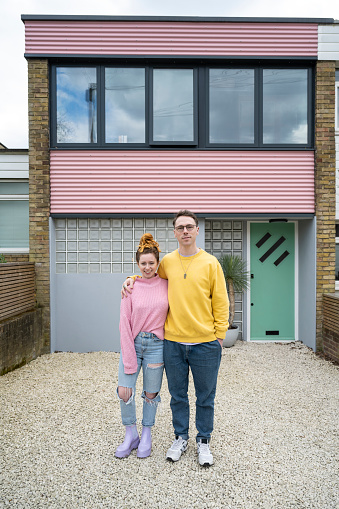 Full length view of proud mid adult homeowners in casual clothing standing with arms around each other and smiling at camera.