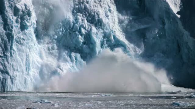 Iceberg begins to crumble throwing spray and waves up in its monstrous wake.
