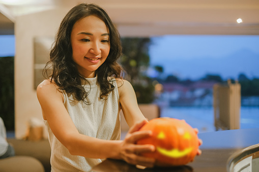 Close-up shot of mid adult Asian woman preparing Halloween decoration above counter table. She's holding a Jack-o'-lantern.