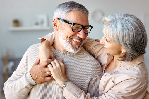 Close up of beautiful smiling senior family couple husband and wife looking at each other with tenderness and love while standing in living room at home, retired man and woman embracing indoors