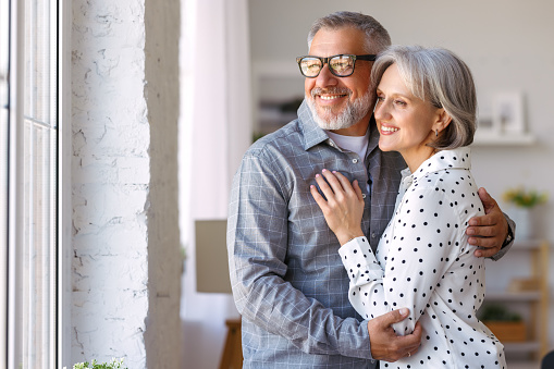 Happy retirement. Smiling beautiful senior caucasian family couple in love embracing while standing near window at home, positive retired wife and husband enjoying retired life together
