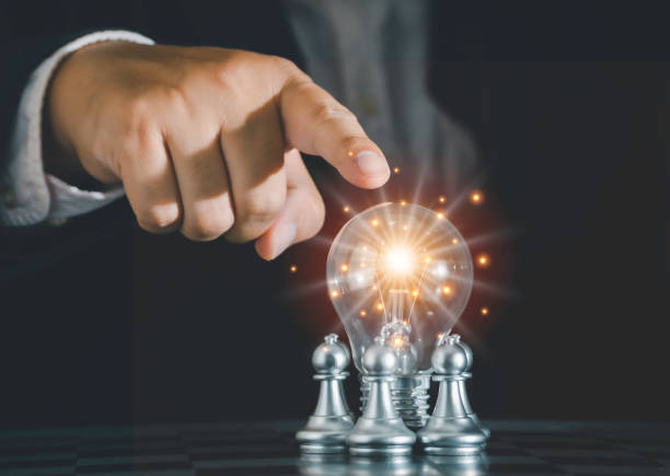 istock Hand of businessman pointing light bulb with fire spark shine glitter is surrounded around silver chess pieces for new idea inspiration fighting with teamwork to victory, strategy concept for success. 1342625747