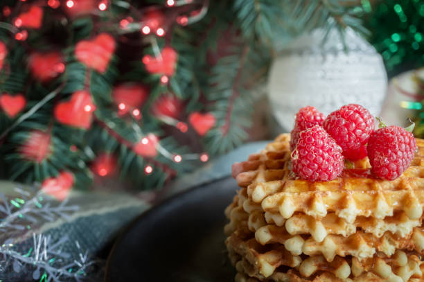 istock Viennese waffles with raspberries and honey on  Christmas table with tree branches and garlands of lights i background. Golden waffles lie on a dark plate against the background of  decorated Christmas tree, with highlights in the shape of hearts 1342627539