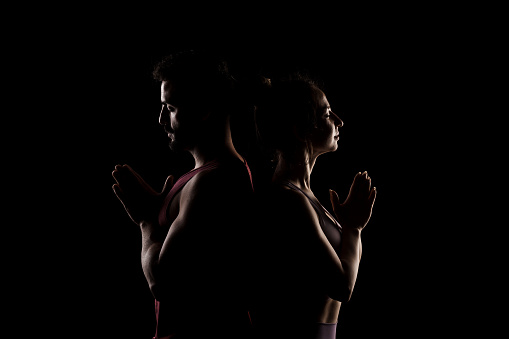 Fit couple posing together. Boy and girl praying back to back side lit silhouettes on black background.