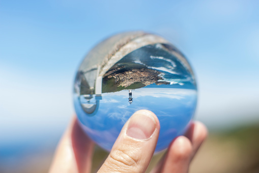 Hand holding a crystal ball reflecting the beautiful and touristic lighthouse of the Galician town of Ribadeo in front of the sea, during a sunny day, concepts, selective focus.