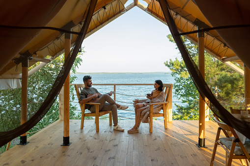 Happy amorous couple relaxing in wooden armchairs on patio by glamping house