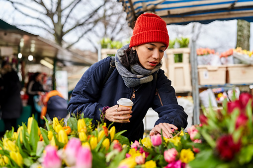 Young woman buying flowers on the local market. Female wearing warm clothing holding a disposable coffee cup looking at the flowers.